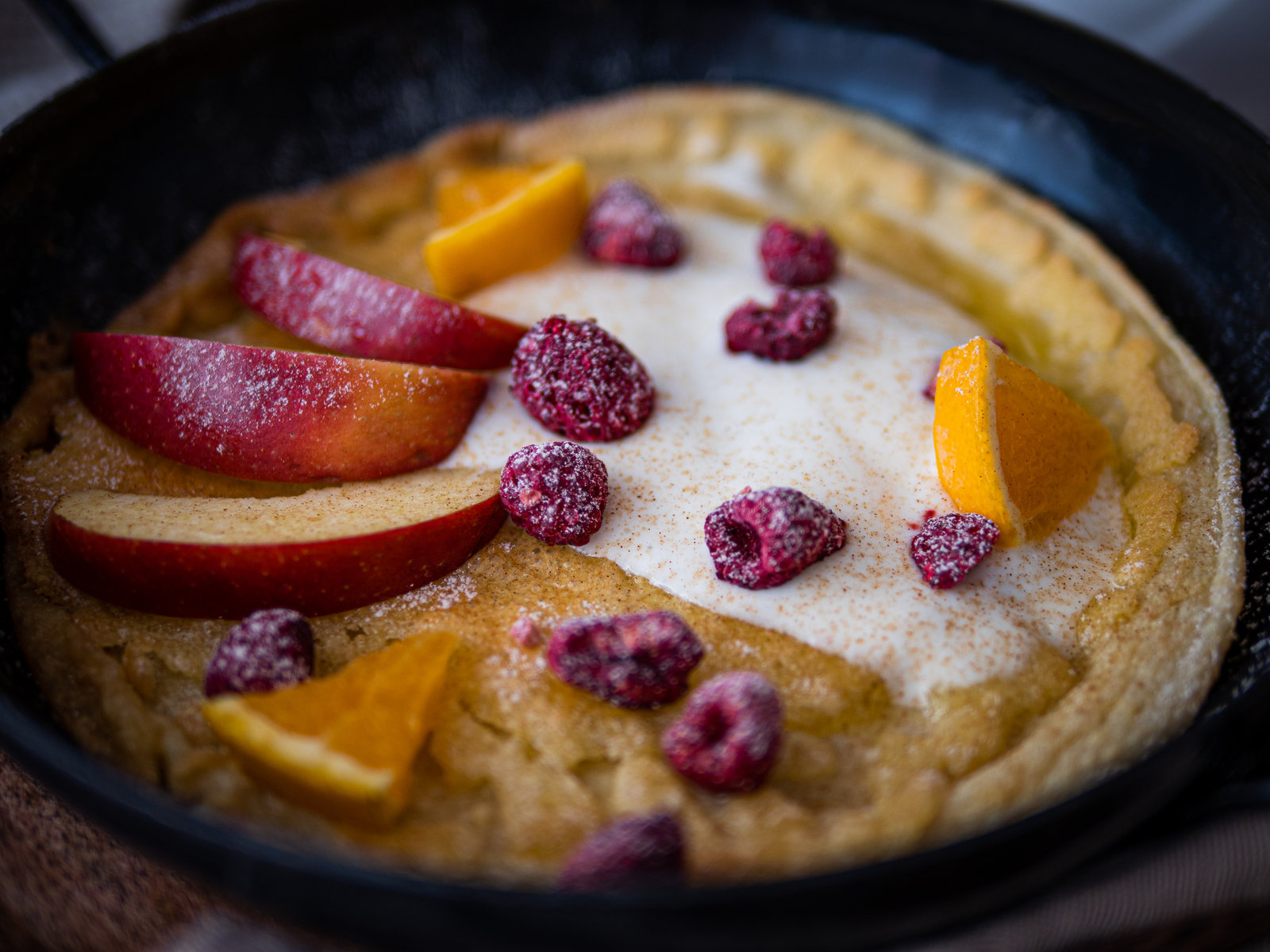 Ein überbackener Pfannkuchen mit Joghurt, Apfelspalten, Orangenstücken und gefriergetrockneten Himbeeren, bestreut mit Zimt-Puderzucker.