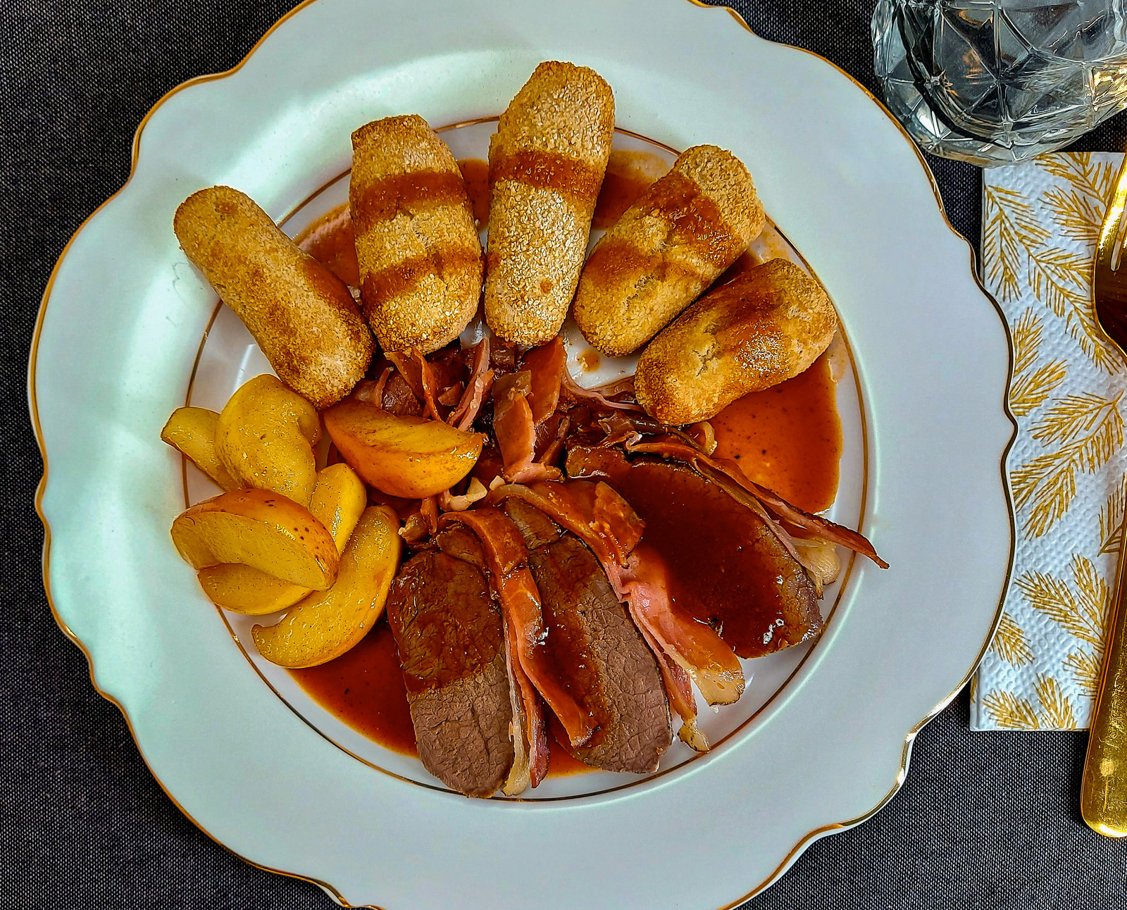 Servierter Braten in Scheiben mit Apfelspalten, Kroketten und Sauce auf einem Teller.