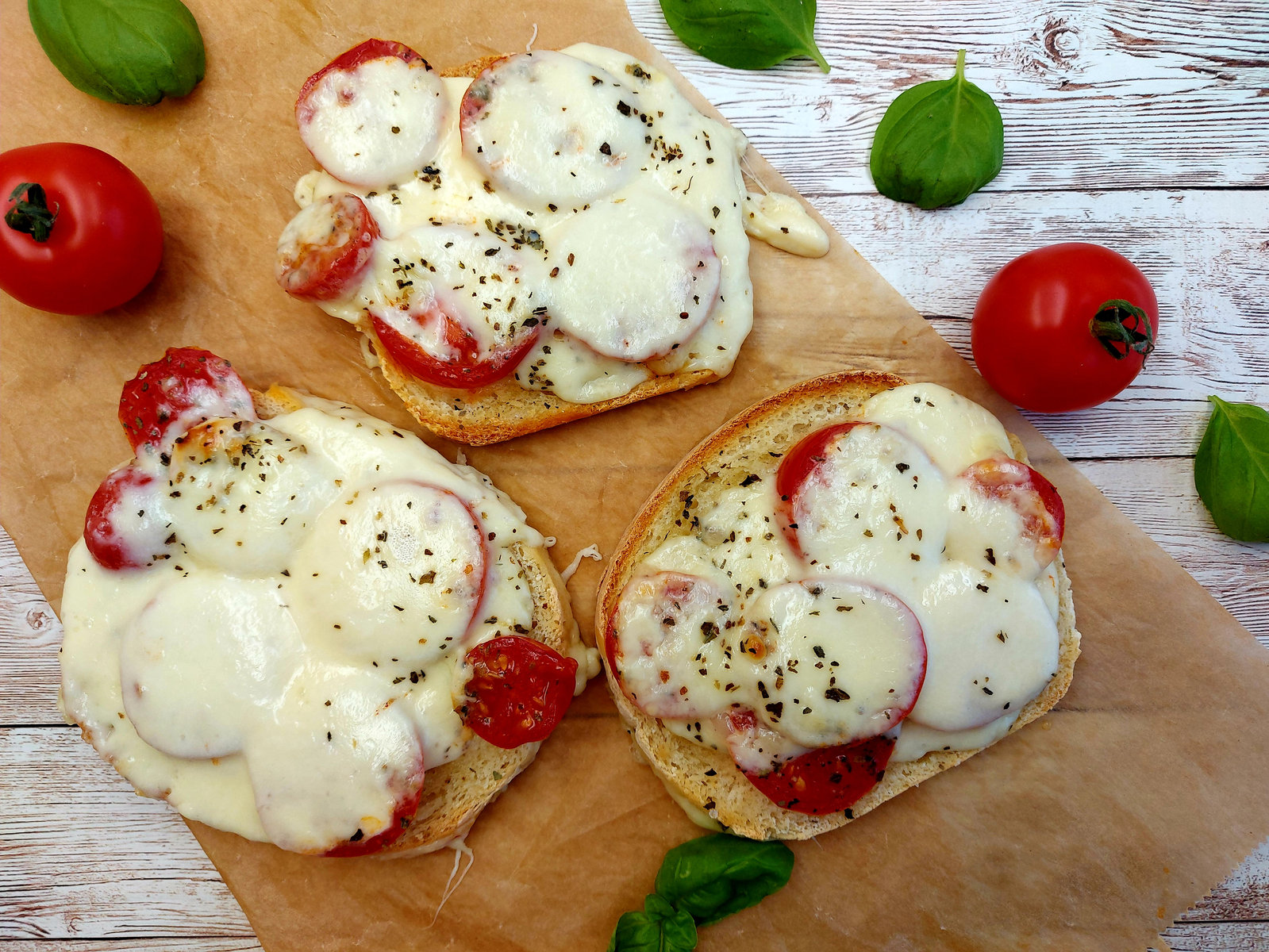 Drei goldbraun gebackene Tomaten Mozzarella Toasts, belegt mit frischem Basilikum, auf einem Holzbrett.