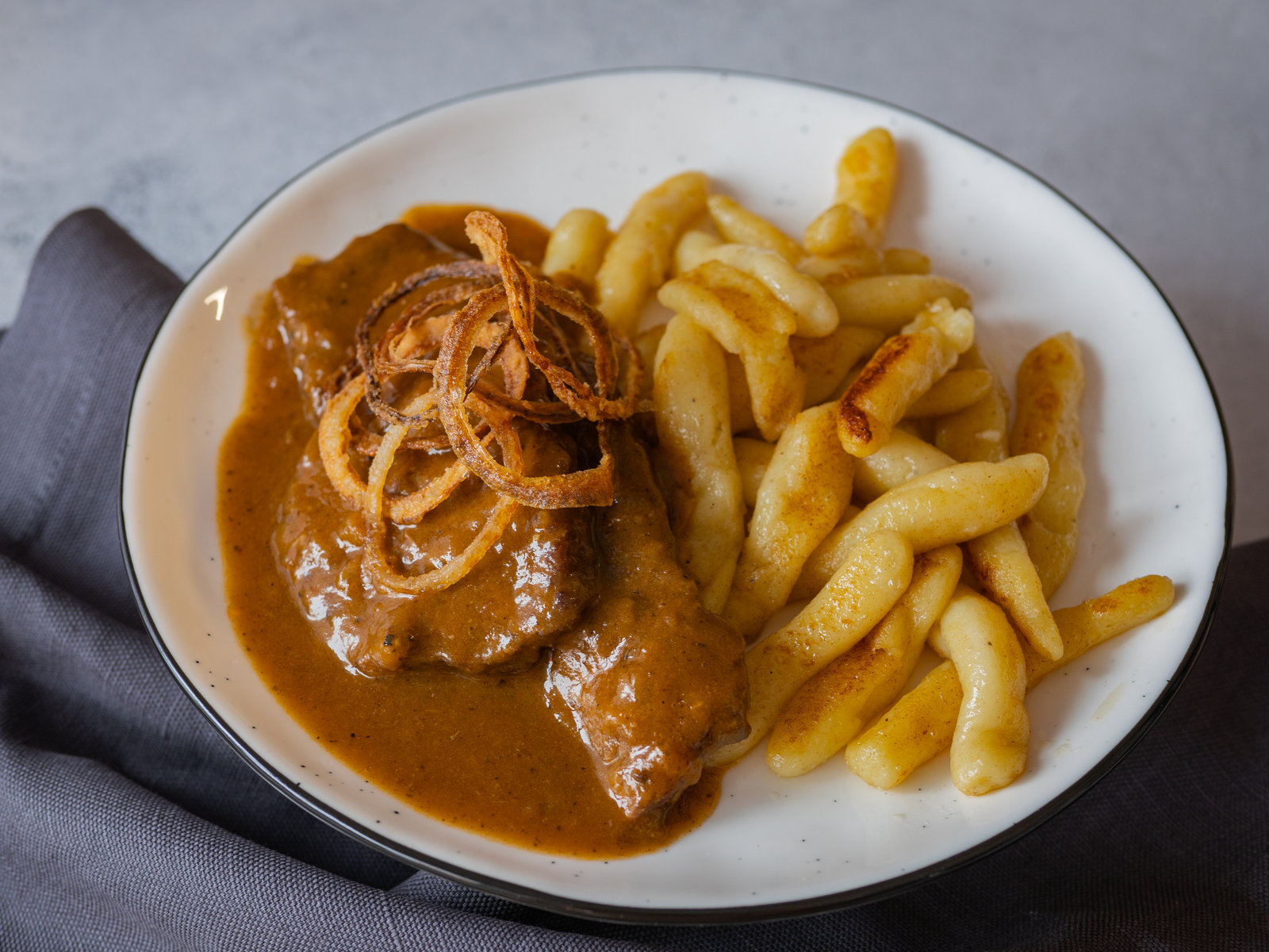 Ein Teller mit Tiroler Zwiebelrostbraten, reichlich Sauce und Röstzwiebeln, dazu Beilagen.