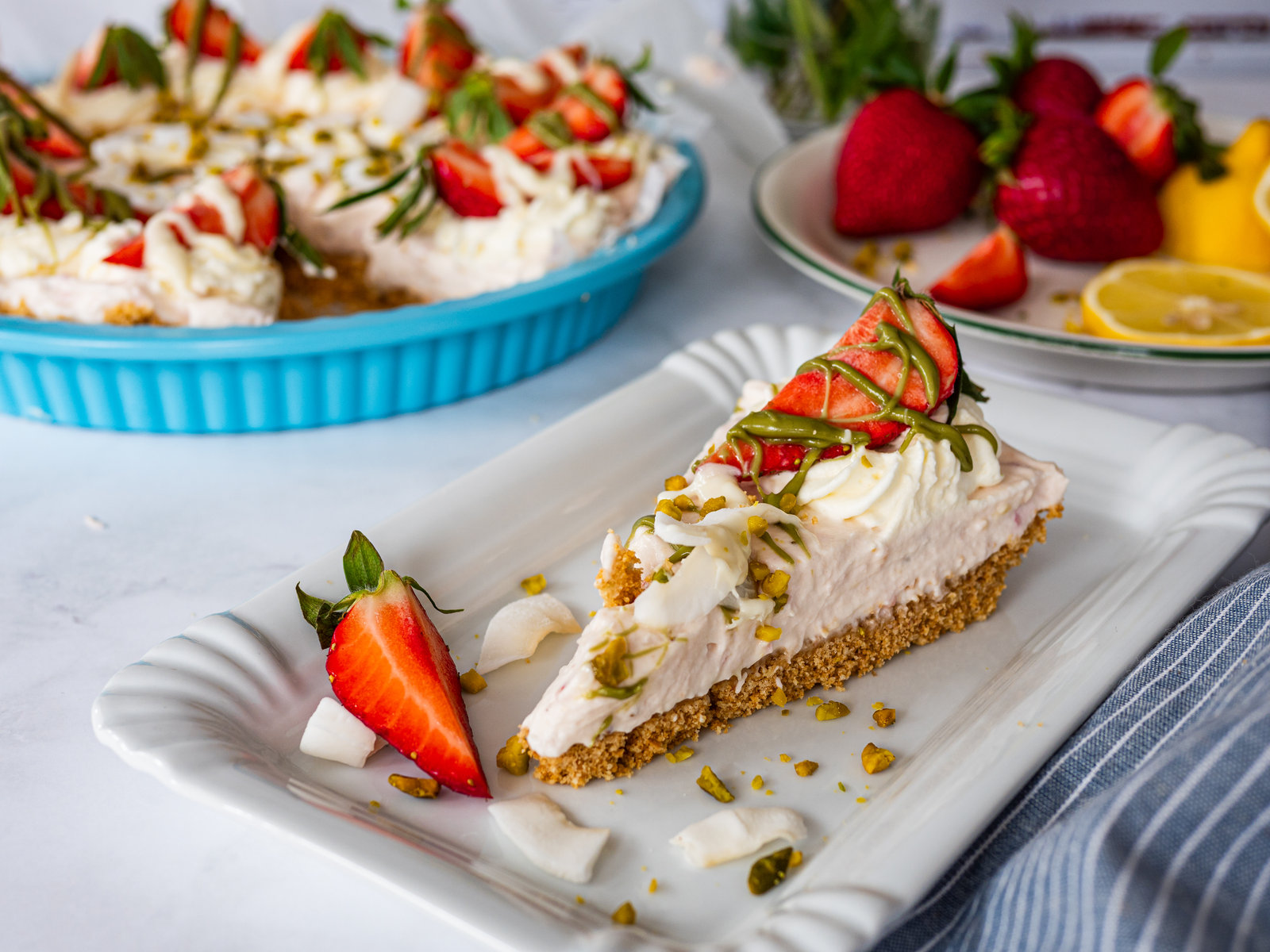Eine angeschnittene No Bake Erdbeer-Tarte mit Keksboden, Erdbeer-Frischkäsecreme und frischen Erdbeeren, verziert mit Pistazien und Kokosstreifen.